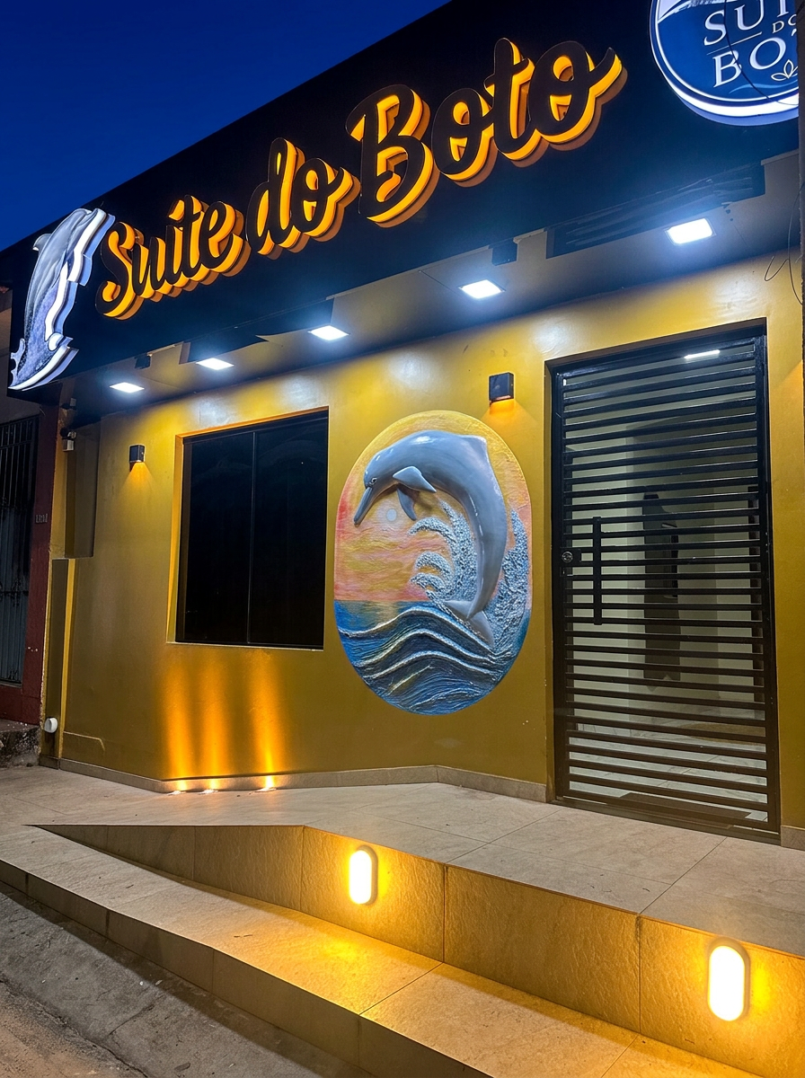Fachada da Suíte do Boto iluminada à noite, com letreiro e painel do boto